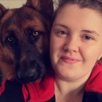 Amy is smiling and wearing a black and red hooded jacket. There is a black and tan german shepherd resting its head on her shoulder.